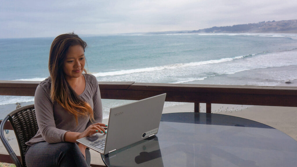 An image of a woman sitting at a table looking at a laptop with the ocean in the background