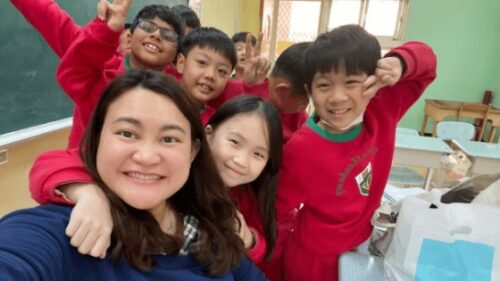 Teacher Shella smiles in a classroom, taking a selfie with her young students.