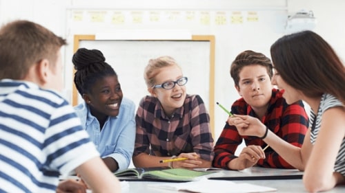 a teacher sitting at a round table with four young teen students who are listening and smiling as she talks and gestures. 