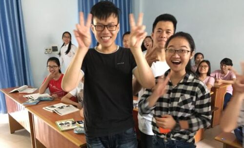 teen students in a classroom, some standing, some sitting, smile and laugh while playing a game.