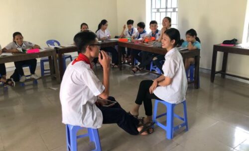 two young adult students practice conversational skills sitting on stools in front of their smiling classmates.