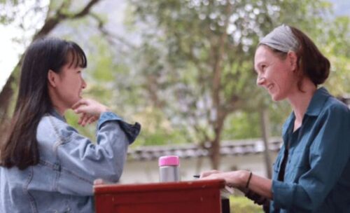 a female teacher and student sit outside at a table smiling and talking.