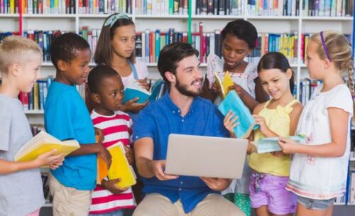 Students gather around a smiling teacher holding a laptop.