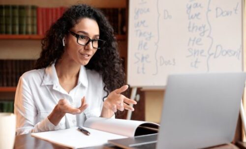 An online teacher talks to students on her laptop with a whiteboard behind her showing vocabulary Do's and Dont's. 