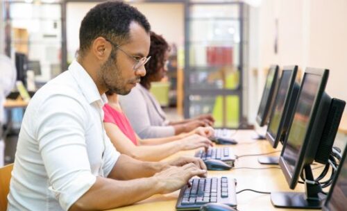 a male teacher sits in the foreground of a long desk with three laptops, as he and two other teachers work on online courses.