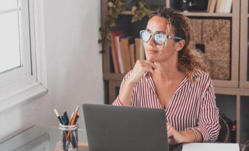 a woman sits at her desk in front of her laptop staring out the window.