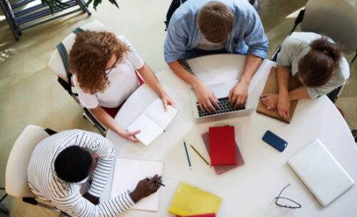 two men and two women sit around a circular table, sharing a laptop and studying.