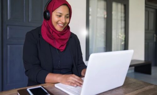 a young woman wearing headphones smiles at her laptop screen while typing.