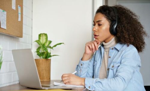 adult female student considers her laptop screen, poised to make notes on a tablet.