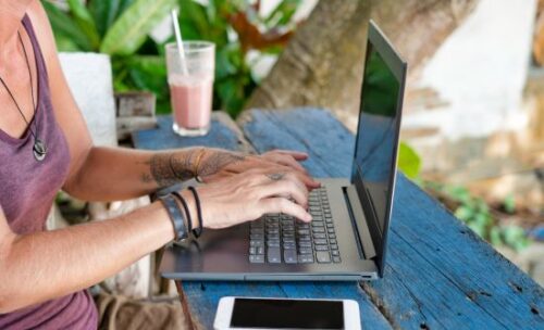 a female teacher types on her laptop sitting on a painted bench in a tropical location.