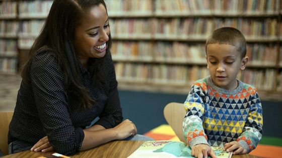 A female English teacher sits at a library table next to a young bog who is pointing to a page in an open book.