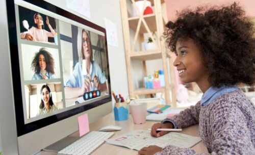 a young student sits at a desk at home videoconferencing with her teacher and other students.