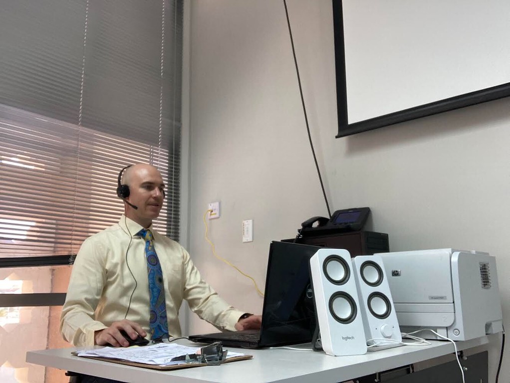 Joshua, a teacher from the US who moved to teach English in Saudi Arabia, gets ready to teach an online class. He wears light clothes, a blue tie and sits in front of a desktop computer.