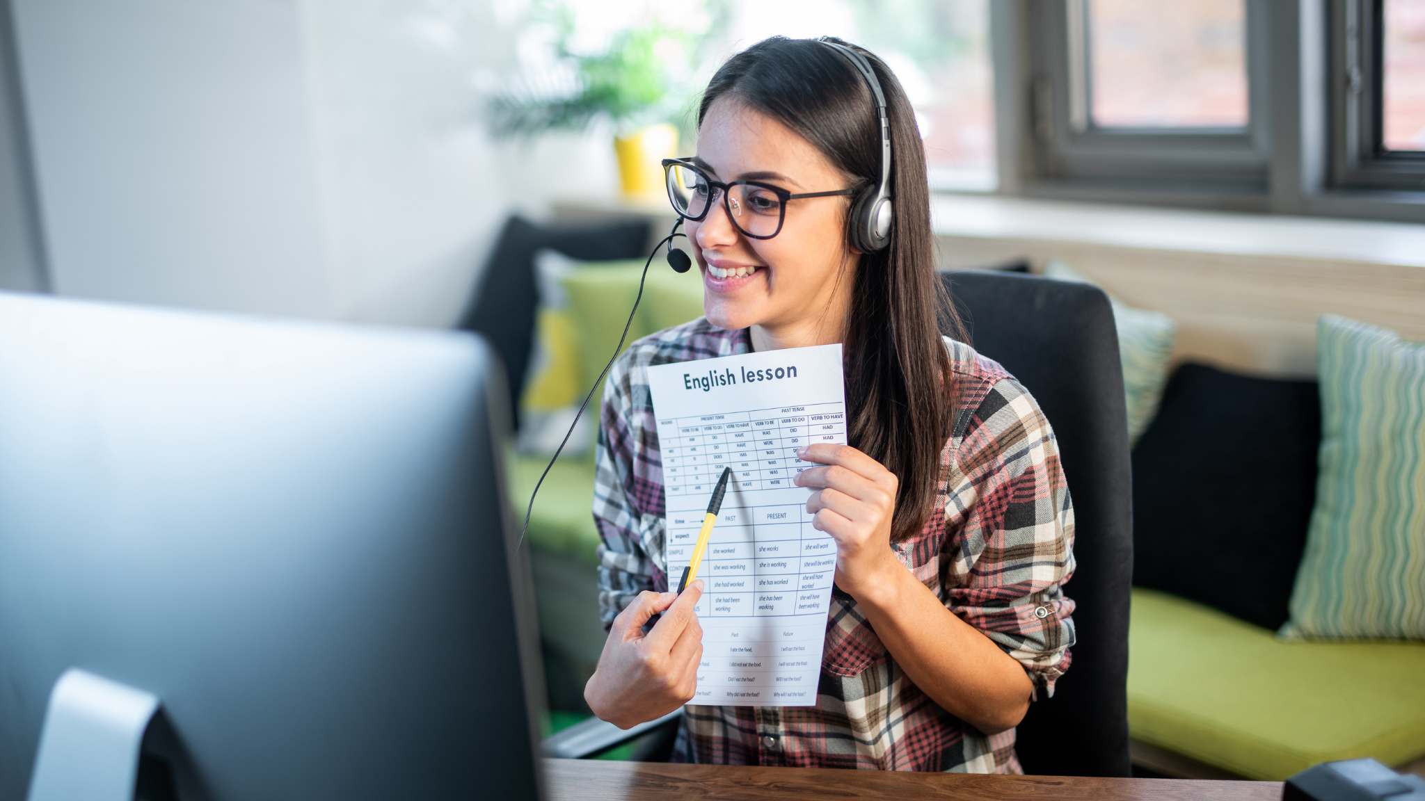 A woman teaches English on her computer while holding up a worksheet.