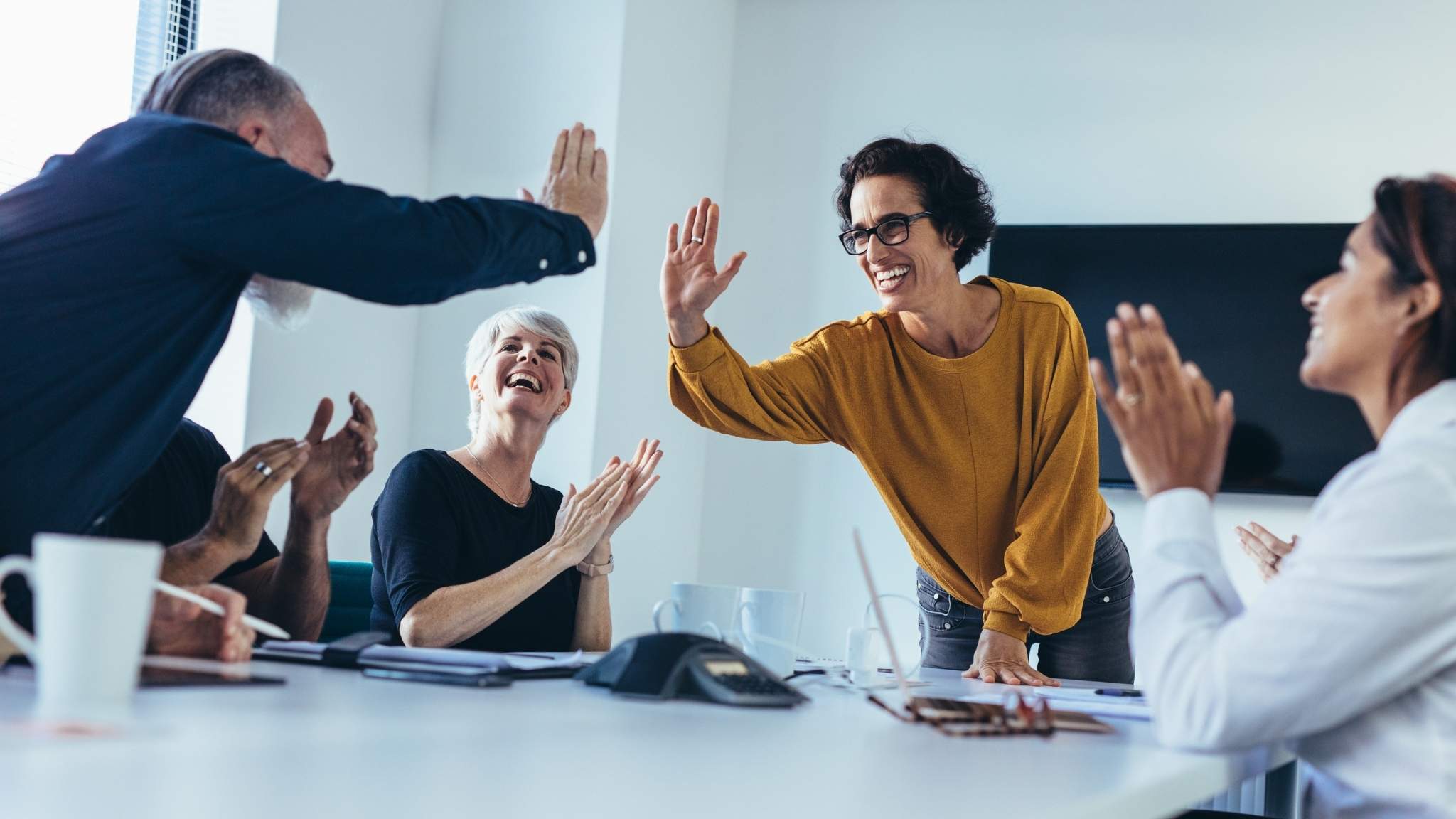 An image of business professionals sitting around a table in a meeting room. Two people are high fiving and the others are clapping happily.