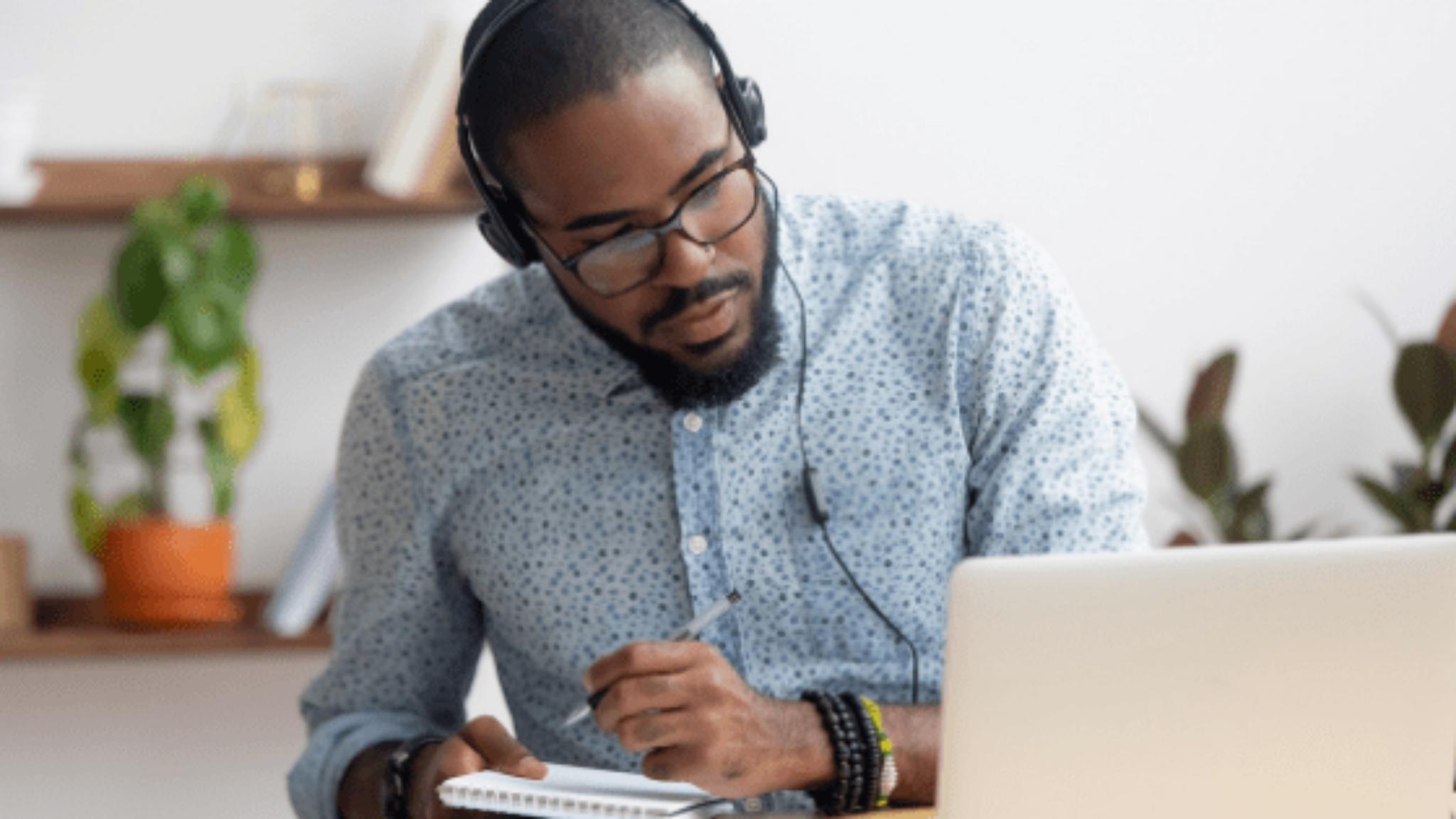 An image of a man looking at a laptop while writing on a notepad and listening to headphones