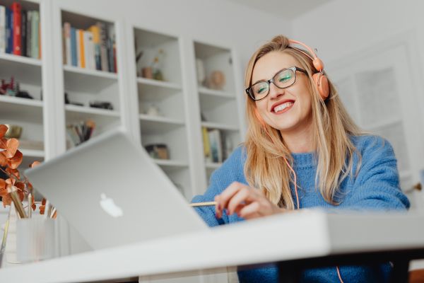 An image of a woman wearing headphones, sitting at a desk in an office and looking at a Macbook