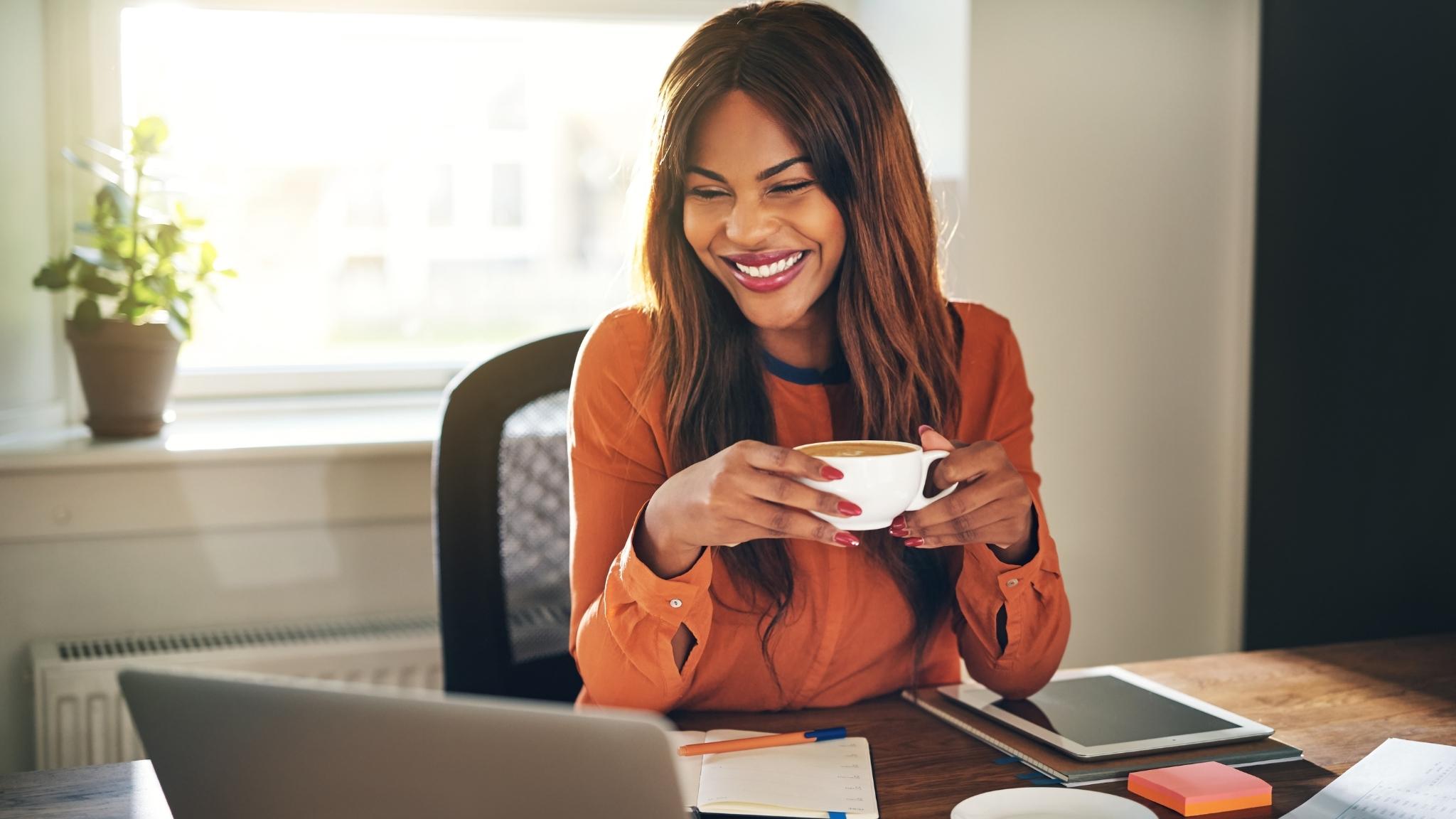 An image of a smiling woman sitting at a desk, holding a cup of coffee, and looking at a laptop