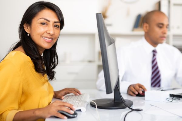 Una imagen de una mujer sentada frente a una computadora, sonriendo a la cámara.