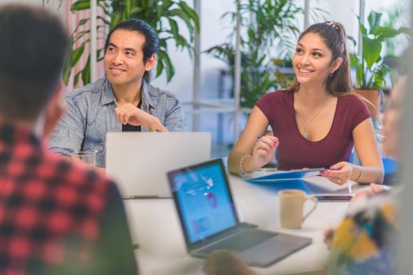 Una imagen de una reunión de equipo donde los miembros del equipo están sentados alrededor de una mesa con sus computadoras portátiles.