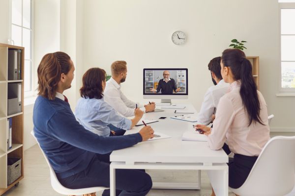 An image of team members sitting around a table while listening to someone attending virtually on a computer