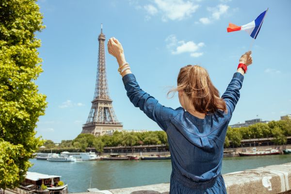 Am image of a woman holding her arms in the air while holding a French flag and looking at the Eiffel Tower