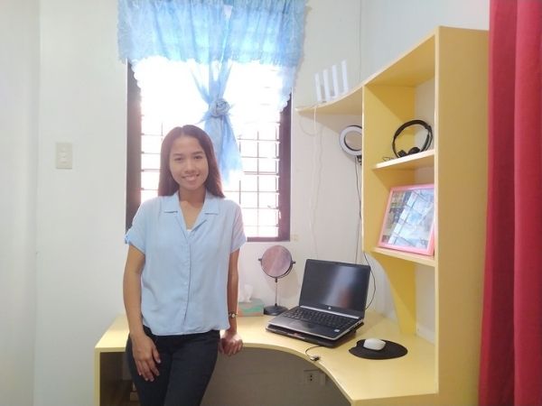 Jersey, an online teacher from the Philippines, standing and smiling beside her home workspace with a laptop and headset on a yellow desk.