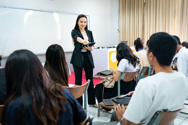 An image of an English teacher at the front of the classroom holding a tablet, giving instructions to students who also have tablets