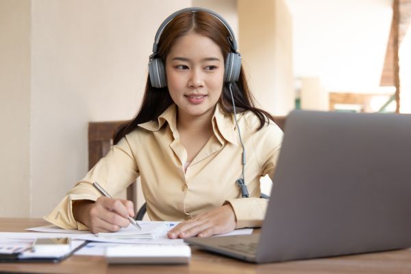 An image of an English student sitting at a desk, wearing headphones, while writing on a notepad