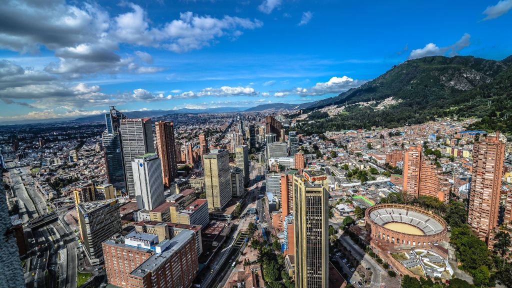 Panoramic view of Bogotá, Colombia, showing modern skyscrapers, residential areas, and the Andes Mountains under a bright blue sky.