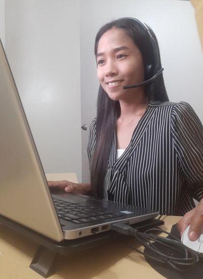 Jersey Cirineo, an online English teacher from the Philippines, smiling while teaching a virtual class from her home workspace using a laptop and headset.