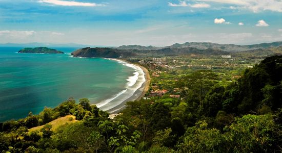 A panoramic view of Playa Jacó in Costa Rica, showing the long curved shoreline with dark sand, turquoise waters, surrounding green hills, and the town along the coast.