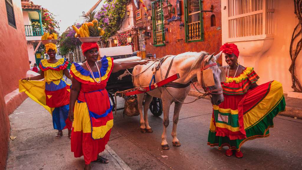 Three women wearing vibrant traditional Colombian dresses stand beside a white horse and carriage on a lively street in Cartagena’s Old City.