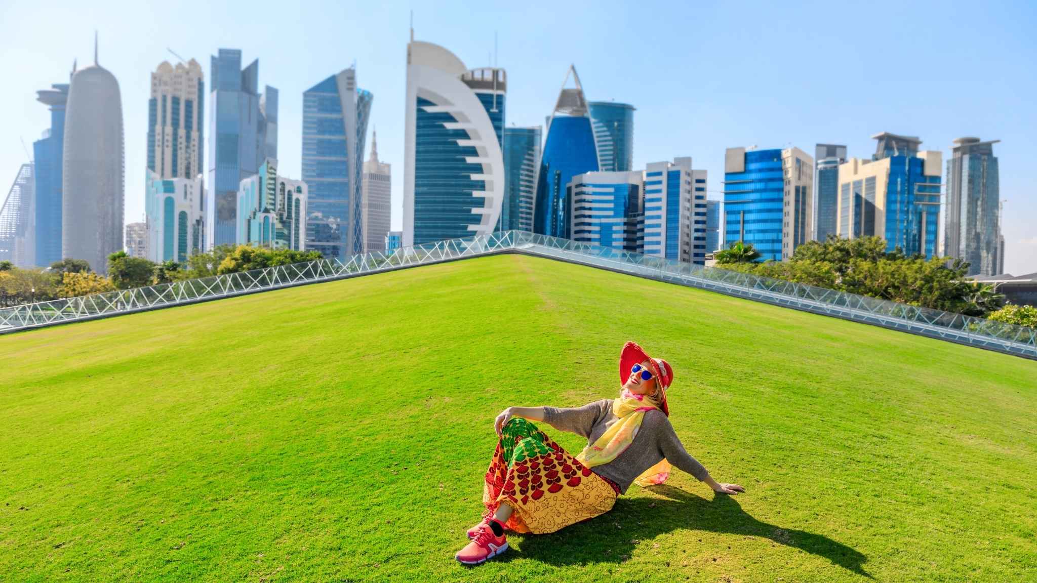 A woman sitting on a green park with Doha's skyline in the back.