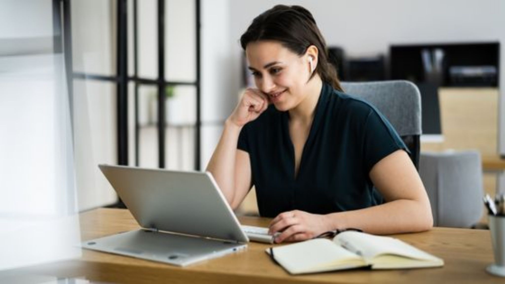 Picture of a woman at her desk, using her laptop with a notebook vby her side.